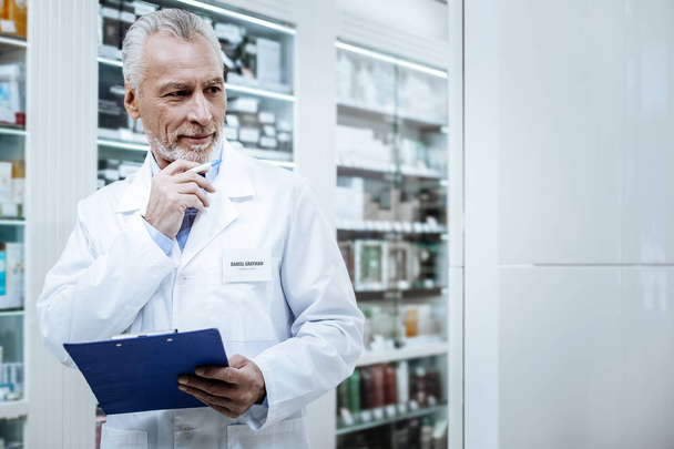Grey-haired bearded pharmacist in a blue shirt comparing medicines - Zdjęcie, obraz
