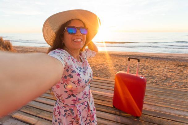 休日、旅行および観光事業コンセプト - ビーチで selfies を取って赤いスーツケースと美しい若い女性 - 写真・画像