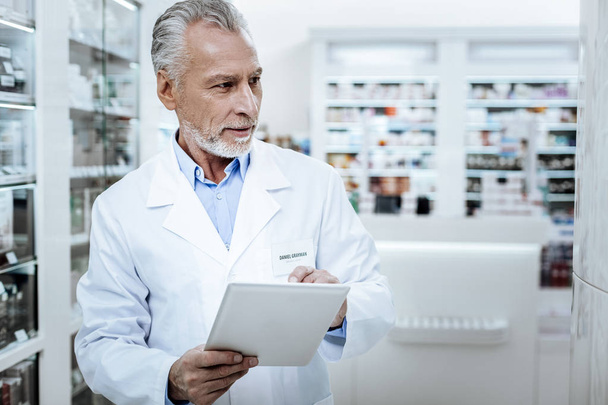 Grey-haired positive pharmacist in a white coat making a revision - Foto, afbeelding