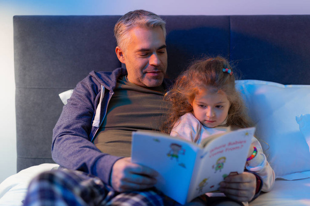 Bebaarde vader lezen van een boek een kleine mooie langharige dochter - Foto, afbeelding
