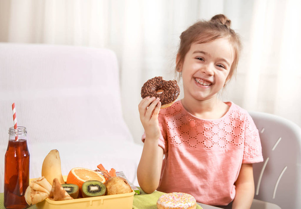 Pieni kaunis iloinen tyttö syö donitsin ja lounaslaatikon, jossa on hedelmiä kotona pöydässä, lasten ruoan käsite.
 - Valokuva, kuva