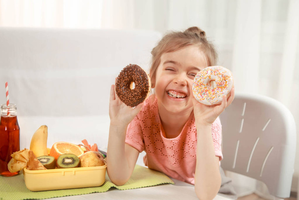 Krásná veselá holčička jíst koblihy a oběd box s ovocem doma u stolu, koncept dětských potravin - Fotografie, Obrázek
