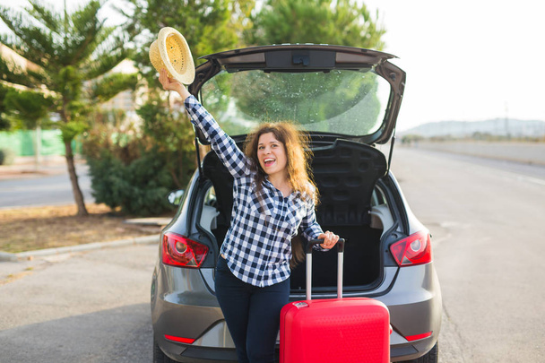 Ταξίδια, διακοπές και άνθρωποι έννοια - νεαρή γυναίκα στέκεται κοντά στο πίσω μέρος του αυτοκινήτου χαμογελώντας και ετοιμάζεται να πάει, κρατώντας το καπέλο - Φωτογραφία, εικόνα