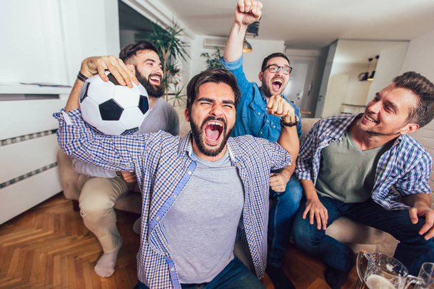 Männer, die zu Hause gemeinsam Sport im Fernsehen verfolgen, schreien fröhlich. Gruppe von Freunden sitzt auf der Couch und schaut sich ein Fußballspiel an. - Foto, Bild