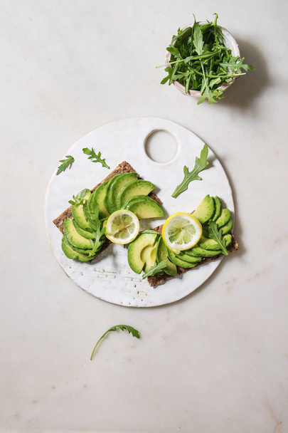 Vegan sandwiches with sliced avocado and lemon on rye bread, arugula salad served on ceramic board over white marble background. Flat lay, space - Photo, image