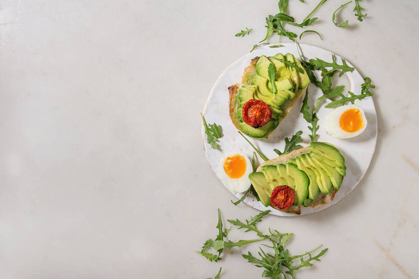 Vegetarian sandwich with sliced avocado, sun dried tomatoes, egg, arugula served on ceramic board over white marble background. Flat lay, space - Photo, image