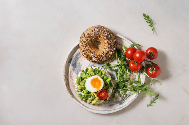Vegetarian whole grain bagel sandwich with chopped avocado, cream cheese, sun dried tomatoes, egg, arugula served on ceramic plate over white marble background. Flat lay, space - Zdjęcie, obraz