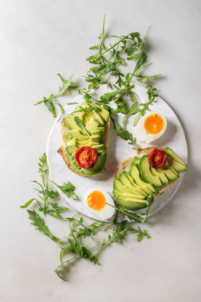 Vegetarian sandwich with sliced avocado, sun dried tomatoes, egg, arugula served on ceramic board over white marble background. Flat lay, space - Fotoğraf, Görsel