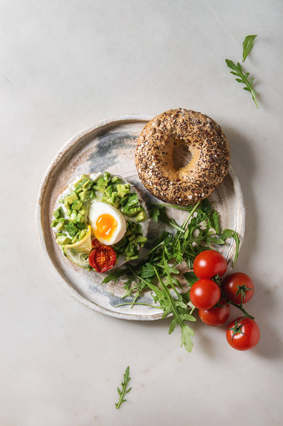 Vegetarian whole grain bagel sandwich with chopped avocado, cream cheese, sun dried tomatoes, egg, arugula served on ceramic plate over white marble background. Flat lay, space - Photo, image