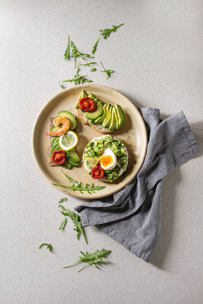 Variety of vegetarian sandwiches with sliced avocado, sun dried tomatoes, egg, shrimps, arugula served on ceramic plate over white grey spotted background. Flat lay, space - Zdjęcie, obraz