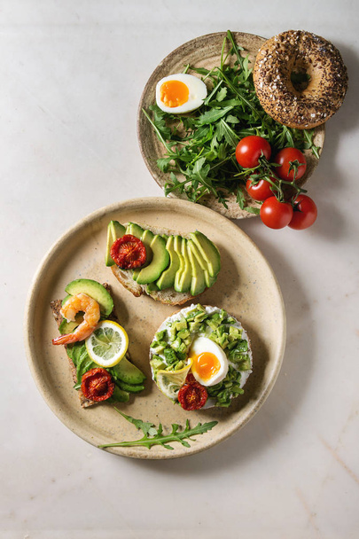 Variety of vegetarian sandwiches with sliced avocado, sun dried tomatoes, egg, shrimps, arugula served on ceramic plate over white grey spotted background. Flat lay, space - Foto, immagini