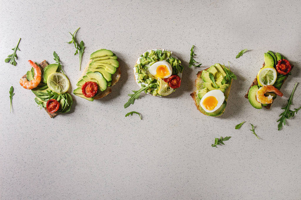Variety of vegetarian sandwiches with sliced avocado, sun dried tomatoes, egg, shrimps, arugula served over white grey spotted background. Flat lay, space - Valokuva, kuva