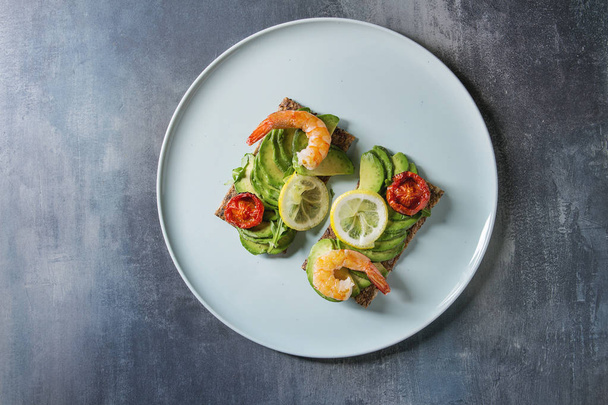 Vegetarian sandwich with sliced avocado, sun dried tomatoes, shrimps, lemon served on ceramic plate over blue texture background. Flat lay, space - Foto, immagini