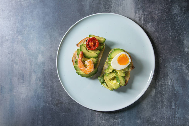 Vegetarian sandwich with sliced avocado, sun dried tomatoes, egg, shrimps, served on ceramic plate over blue texture background. Flat lay, space - Φωτογραφία, εικόνα