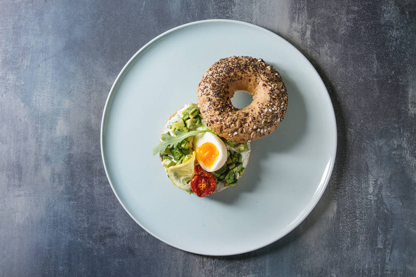 Vegetarian whole grain bagel sandwich with chopped avocado, cream cheese, sun dried tomatoes, egg, served on ceramic plate over blue texture background. Flat lay, space - Foto, Imagem