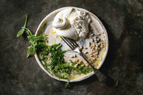 Sliced Italian burrata cheese, fresh arugula salad, pine nuts and olive oil in white ceramic plate over dark metal background. Flat lay, space - Foto, immagini