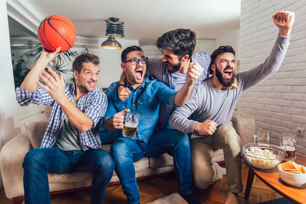 Счастливые друзья или любители баскетбола, которые смотрят баскетбольную игру по телевизору и празднуют победу дома.
. - Фото, изображение