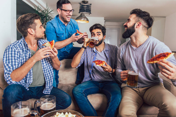 Skupina čtyř mužských přátel pije pivo a doma jí pizzu. - Fotografie, Obrázek