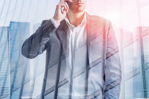 Portrait of unrecognizable young businessman wearing gray suit and talking on smartphone over cityscape background with double exposure of skyscraper. Toned image - Photo, image
