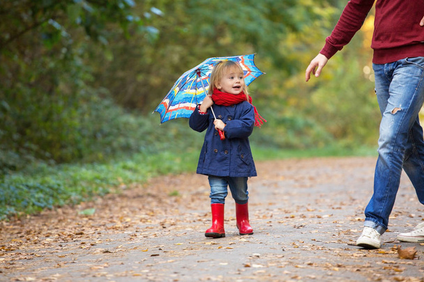 A small girl dressed in rubber boots and raincoats, holds an umbrella, laughs and runs through an autumn park. - 写真・画像