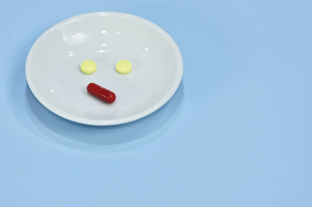 Sad face made of medicine. pain medication tablets. colored pills on porcelain dish. Assorted pharmaceutical pills, tablets and capsules over blue background. - Photo, Image