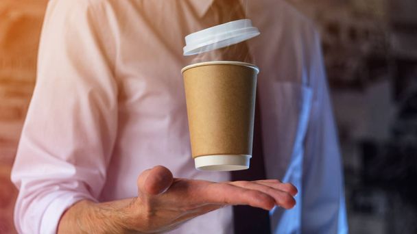 Barista in a white shirt with a tie holds floating in the air a paper cup with a hot drink on his outstretched arms. Barista wizard concept. Background for advertising and placing logo on your drink - Foto, Imagen
