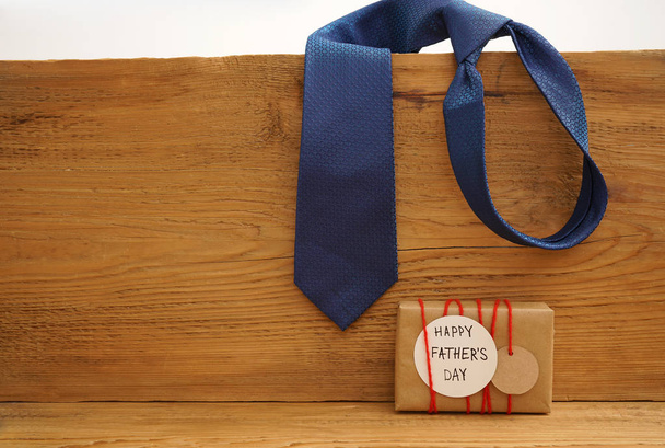 craft paper wrapped gift box with craft paper tag card on wooden background, top view,  Father's Day concept. tie hanging  on wooden plank.  - Fotografie, Obrázek