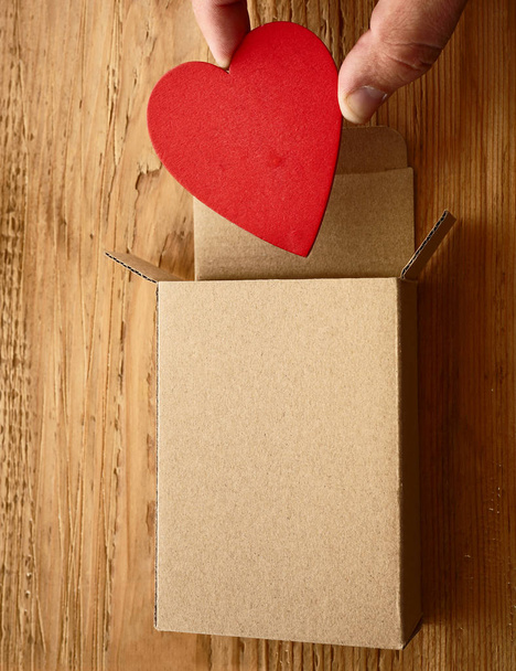 Мужская рука держит одно красное сердце над открытой коробкой подарков на деревянном фоне. С Днем Святого Валентина. 14 февраля
.    - Фото, изображение