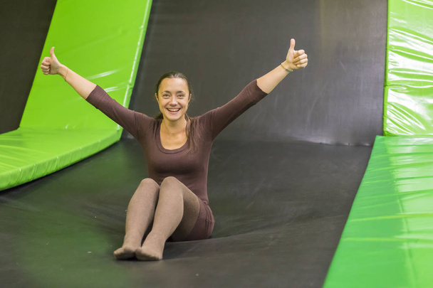 jovem mulher em vestido de ajuste marrom no trampolim no centro de trampolim. foto vertical. Menina feliz no trampolim. Jovem feliz no trampolim
 - Foto, Imagem