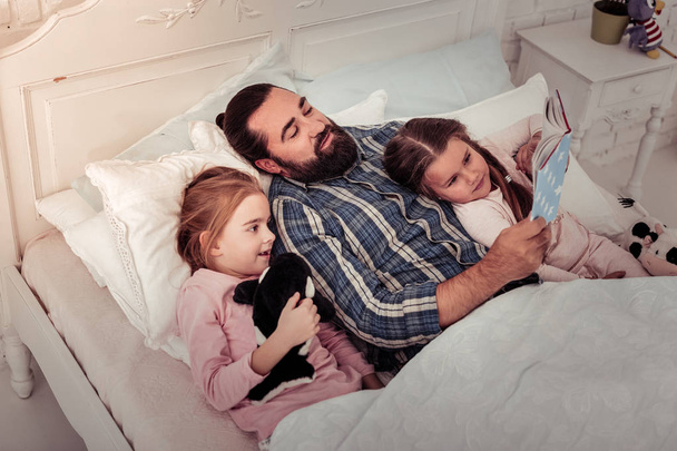 Care and love. Nice loving father lying with his daughters while reading them a book - Fotó, kép