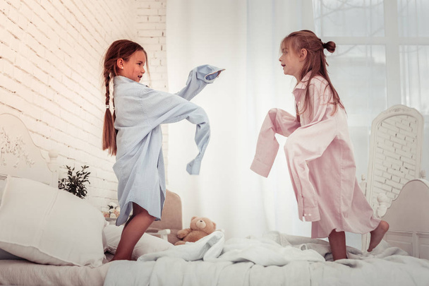 Dads clothes. Joyful nice happy girls wearing male shirts while jumping on the bed - Foto, Bild
