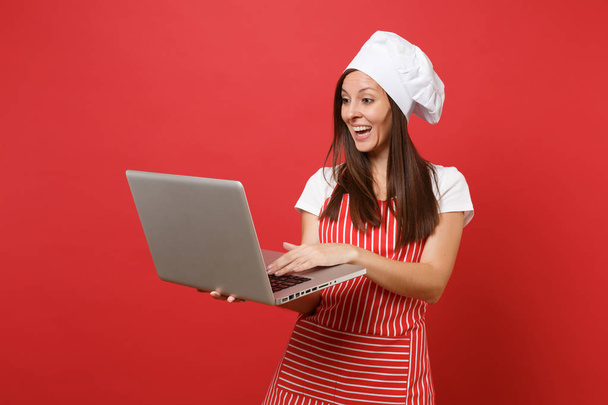 Домогосподарка кухарки кухарки або пекарні в смугастій фартусі, біла футболка, шапка кухарів ізольовані на червоній стіні. Весела домогосподарка шукає рецепт на ноутбуці. Зробити концепцію простору копіювання
 - Фото, зображення