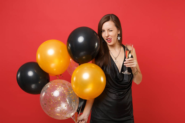 Mujer sexy en vestido negro celebrando labios lamiendo parpadeo, sosteniendo una copa de champán, globos aéreos aislados sobre fondo rojo. Día de San Valentín, Feliz Año Nuevo concepto de fiesta de fiesta maqueta cumpleaños
 - Foto, Imagen