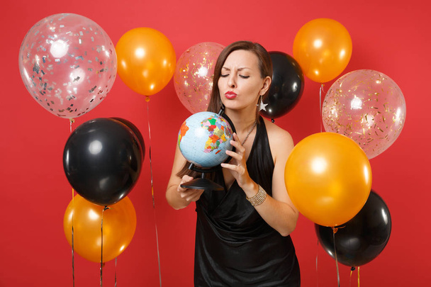 Озадаченная молодая женщина в маленьком черном платье празднует, глядя на мир в руках на красном фоне воздушных шаров. International Women 's Day, Happy New Year, birthday mockup holiday party concept
 - Фото, изображение