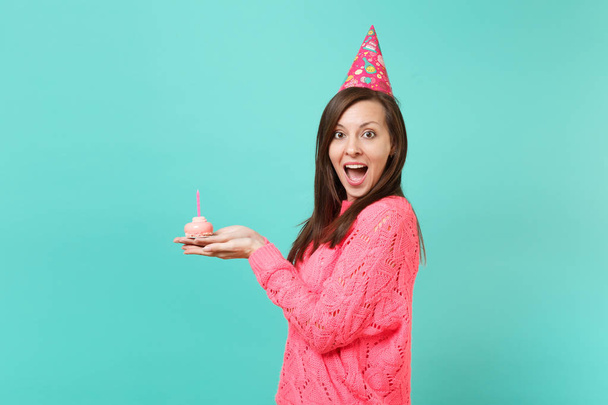 ニットのピンクのセーター、口の中を維持する誕生日帽子に興奮した女性の側面図を開き、青色の背景に分離されたキャンドルとケーキを保持見てみるとビックリ。人々 のライフ スタイルのコンセプトです。コピー スペースをモックアップします。 - 写真・画像
