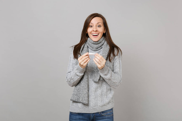 Χαρούμενη ενθουσιασμένος νεαρή γυναίκα στο κασκόλ γκρι πουλόβερ κρατώντας θερμόμετρο που απομονώνονται σε φόντο γκρίζο τοίχο στο studio. Υγιεινού τρόπου ζωής, άρρωστο άρρωστο νόσου θεραπεία, το κρύο σεζόν έννοια. Κοροϊδεύετε αντίγραφο χώρου - Φωτογραφία, εικόνα