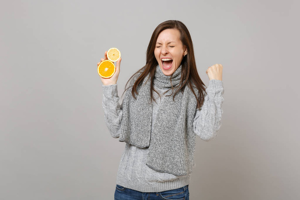 Szczęśliwa młoda kobieta w szary sweter, szalik, krzyczy, zaciskając pięści jak zwycięzca przytrzymaj cytryny pomarańczowy na białym tle na szarym tle. Moda na zdrowy styl życia ludzi szczere emocje zimowym koncepcja - Zdjęcie, obraz