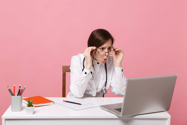 Tired sad female doctor sits at desk work on computer with medical document in hospital isolated on pastel pink wall background. Woman in medical gown glasses stethoscope. Healthcare medicine concept - Φωτογραφία, εικόνα