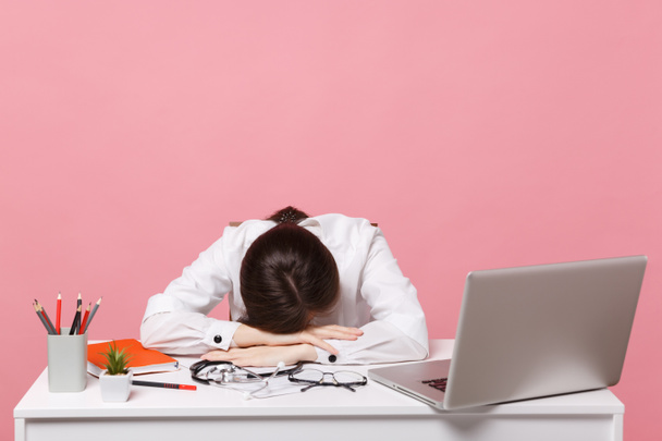 Female doctor laid her head down on desk, work on computer, medical document in hospital isolated on pastel pink background. Woman in medical gown glasses stethoscope. Healthcare medicine concept - Photo, Image
