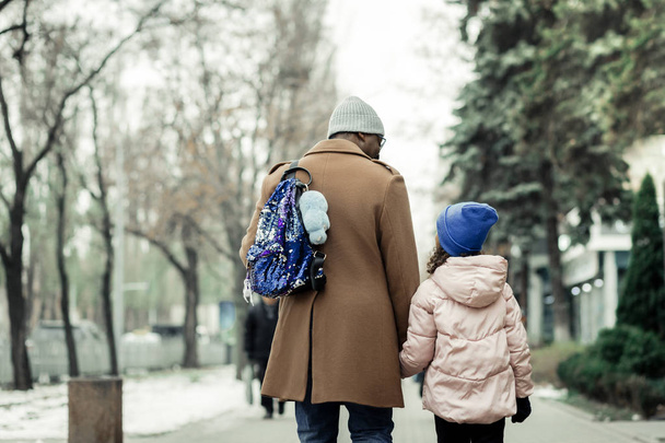 Loving father wearing brown coat caring backpack of daughter - Fotoğraf, Görsel