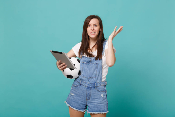 Зачарована молода жінка-футболістка тримає футбольний м'яч, використовуючи планшетний комп'ютер, розтягуючи руки ізольовано на синьому бірюзовому фоні стіни. Люди емоції, спортивна сімейна концепція способу життя
 - Фото, зображення