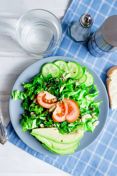  Fresh  green salad with cucumber, lettuce, avocado and tomatoes and glass water on a white wooden table. Healthy food lifestyle concept. Flat lay. Copy space for text - Foto, afbeelding