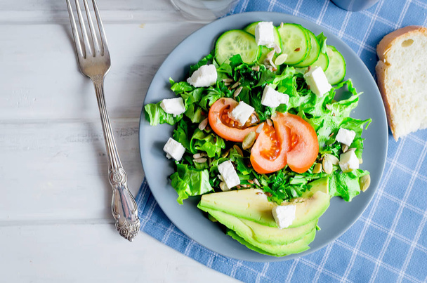  Fresh  green salad with cucumber, lettuce, avocado, tomatoes and soft cheese and glass water on the white wooden table. Healthy food lifestyle concept. Flat lay. Copy space for text - Foto, afbeelding