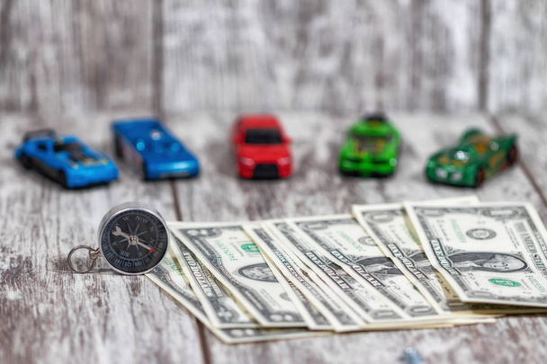 Cash in dollars, a small compass and colorful toy cars lie on a light wooden table - Fotografie, Obrázek