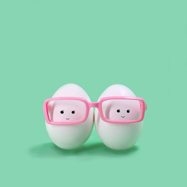 Due uova in bicchieri rosa come gemelli siamesi. Personaggio divertente in stile kawaii. Decorazione creativa pasquale. Concetto pasquale minimo
 - Foto, immagini