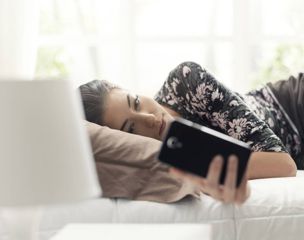 Young beautiful woman relaxing on the bed and connecting with her smartphone, she is chatting with her friends and reading messages - Valokuva, kuva