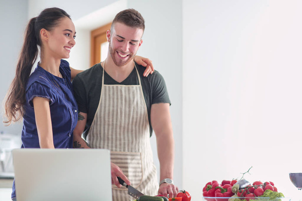 Young man cutting vegetables and woman standing with laptop in the kitchen - Foto, imagen