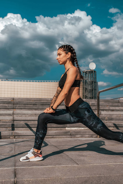 Girl athlete summer city. Makes a warm-up, warms up before playing sports. In leggings sneakers. Woman with tattoos. Before jogging run. - Foto, imagen