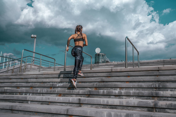 Girl athlete, summer city. Listening to music with headphones, morning jogging on the stairs jumps. In leggings sneakers. Woman with tattoos. In hands of a smartphone. Free space for text. - Фото, изображение
