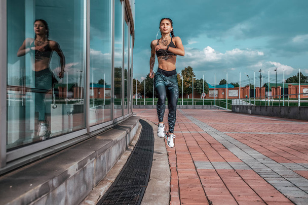 Girl athlete, summer city. Listens to music in headphones, jumps on the evening run. In leggings sneakers. Woman with tattoos. Free space for text. In hands of a smartphone. - Foto, afbeelding